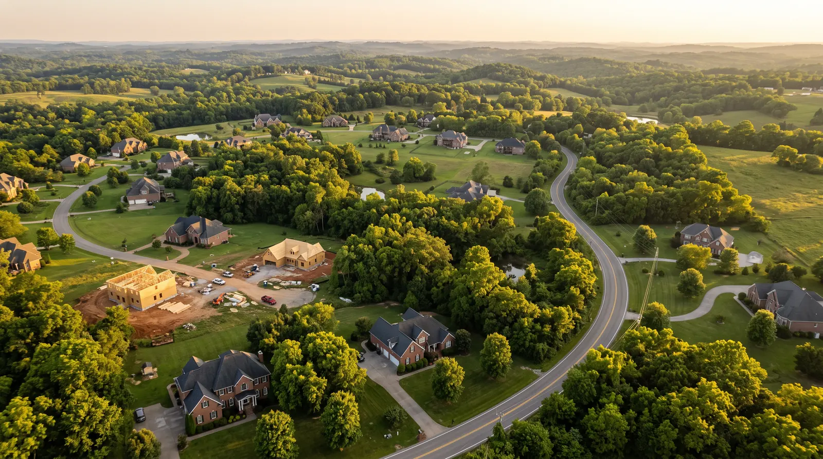 Aerial View of Middle Tennessee Neighborhoods | Music City Builder Group An aerial view over a green Middle Tennessee area at golden hour showing brick and stone homes, a few homes under construction, mature trees and rolling hills with a winding road