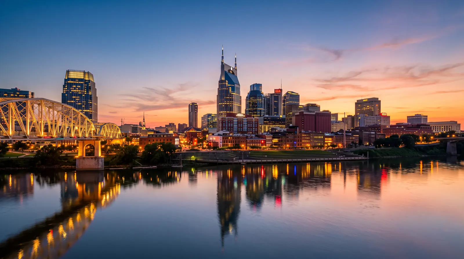 The Nashville skyline glowing at blue-hour dusk beyond the Cumberland River with a calm reflection, a warm sunset gradient in the sky and the lit pedestrian bridge